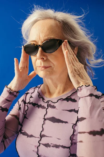 Портрет старшої жінки, що регулює сонцезахисні окуляри ізольовані на синьому — стокове фото