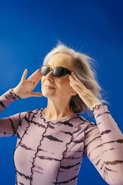 Portrait of satisfied senior woman adjusting sunglasses while looking away isolated on blue — Stock Photo