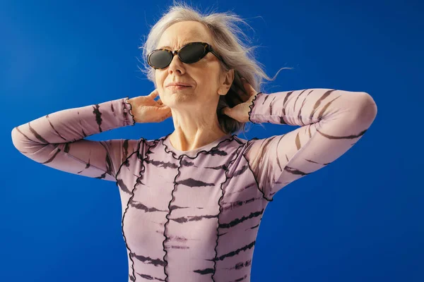 Satisfied elderly woman in sunglasses looking away while posing isolated on blue — Stock Photo