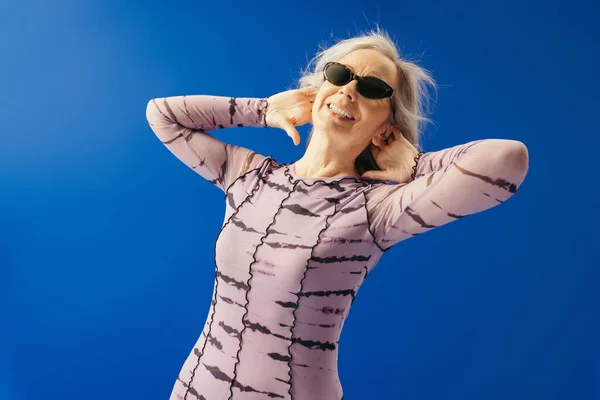 Щаслива літня жінка в одязі і сонцезахисних окулярах посміхається ізольовано на синьому — стокове фото