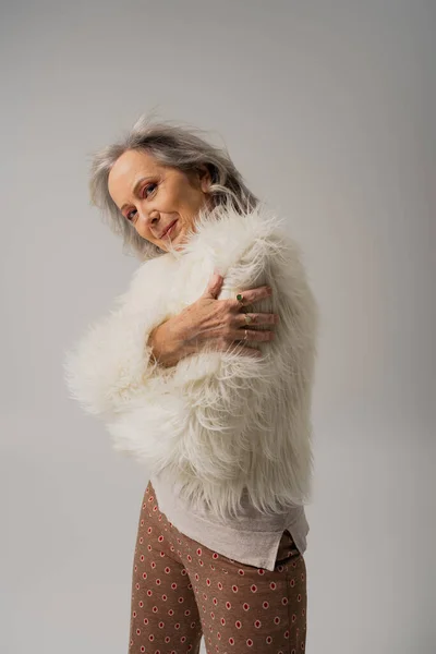 Happy elderly woman in white faux fur jacket posing isolated on grey — Stock Photo