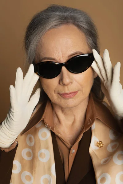 Портрет пожилой женщины в белых перчатках, регулирующей солнцезащитные очки, изолированные на бежевом — стоковое фото