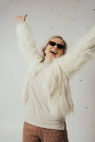 Happy elderly woman in white faux fur jacket and sunglasses raising hands near falling confetti on grey — Stock Photo