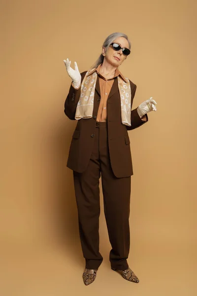 Full length of senior woman in brown formal wear and white gloves standing on beige — Stock Photo