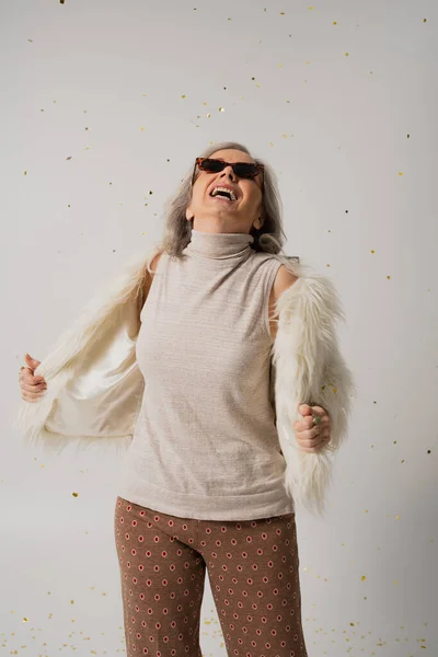 Happy elderly woman in white faux fur jacket and sunglasses looking at falling confetti on grey background — Stock Photo