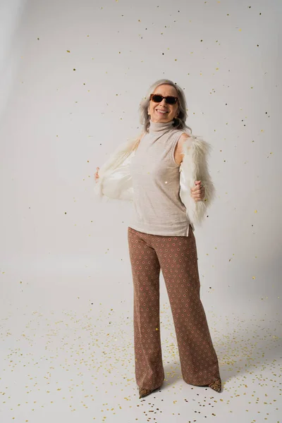Full length of positive elderly woman in white faux fur jacket and sunglasses standing near falling confetti on grey background — Stock Photo