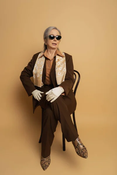 Полная длина пожилой женщины в коричневой формальной одежде и солнечных очках, сидящих на деревянном стуле на бежевом — стоковое фото
