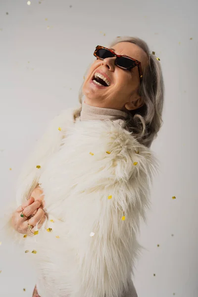 Счастливая пожилая женщина в белом искусственном меховом пиджаке и модных солнцезащитных очках смеялась над падающими конфетти на серый — стоковое фото