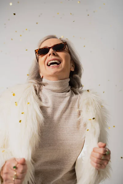 Positive elderly woman in white faux fur jacket and trendy sunglasses laughing near falling confetti on grey background — Stock Photo
