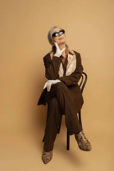 Comprimento total da mulher sênior pensativo no desgaste formal marrom e óculos de sol sentados em cadeira de madeira no bege — Fotografia de Stock