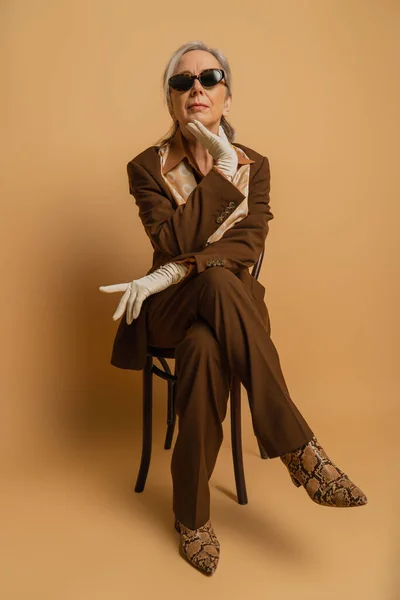 Full length of senior woman in brown formal wear and sunglasses sitting on chair and looking at camera on beige — Photo de stock
