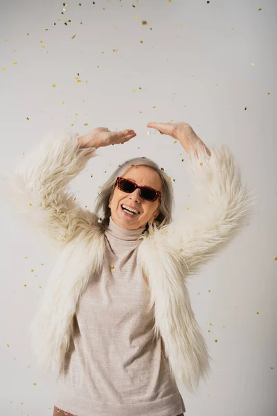 Happy elderly woman in white faux fur jacket and sunglasses standing with raised hands near falling confetti on grey — Stock Photo