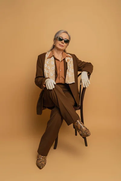 Full length of elderly woman in brown formal wear and sunglasses sitting on chair and looking at camera on beige — Stock Photo
