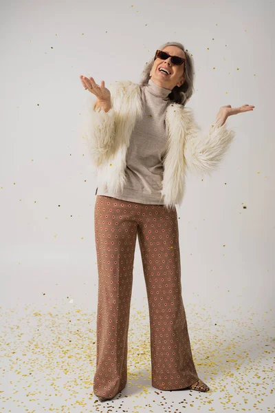 Longitud completa de feliz anciana en chaqueta de piel sintética blanca y gafas de sol de moda sonriendo cerca de caer confeti en gris — Stock Photo