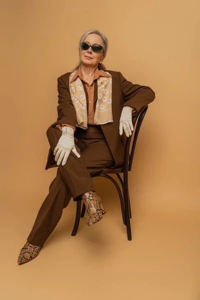 Pleine longueur de femme âgée en costume marron et gants assis sur la chaise et regardant la caméra sur beige — Photo de stock