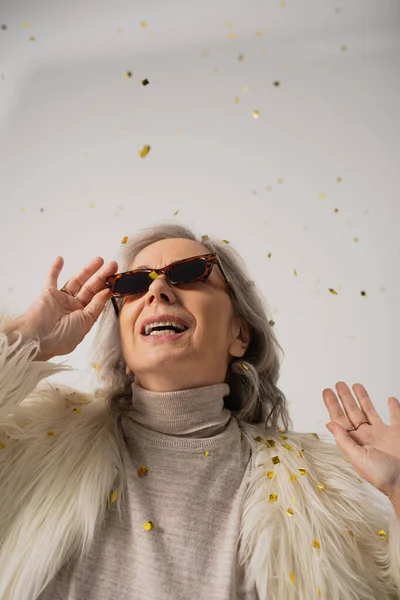 Happy senior woman in white faux fur jacket and trendy sunglasses smiling near falling confetti on grey background — Stock Photo