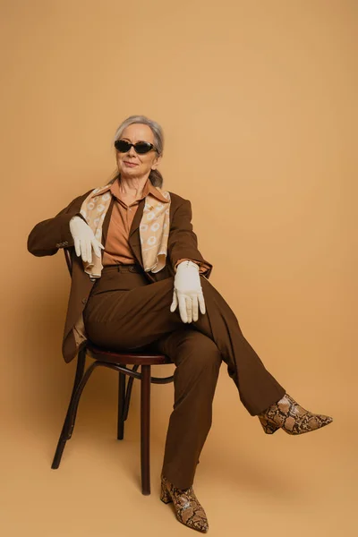 Полная длина веселой пожилой женщины в коричневом костюме и солнцезащитных очках, сидящих на стуле и смотрящих в камеру на бежевом — стоковое фото