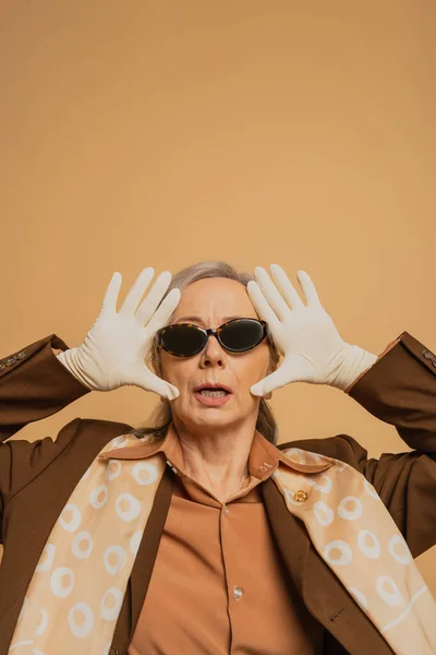 Surprised senior woman in sunglasses and suit posing isolated on beige — Stock Photo