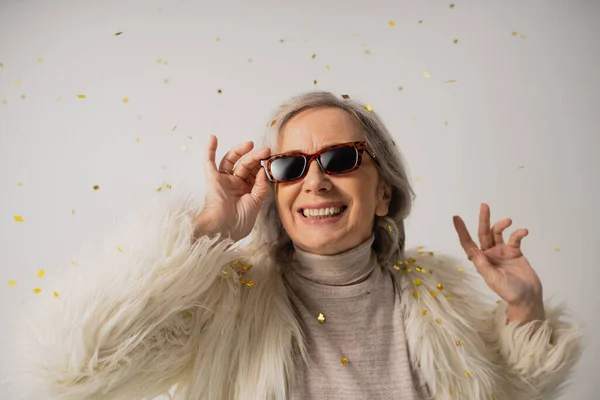 Cheerful senior woman in white faux fur jacket adjusting trendy sunglasses near falling confetti on grey background — Stock Photo