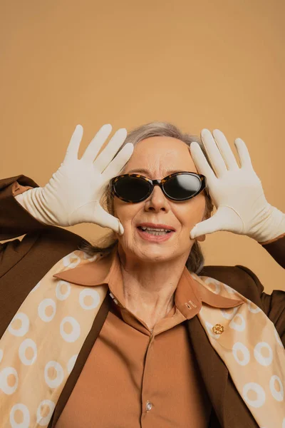 Heureuse et élégante femme âgée en lunettes de soleil et gants blancs posant isolé sur beige — Photo de stock