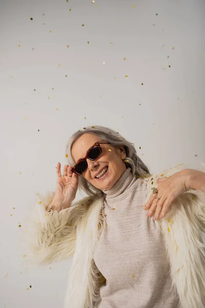 Mulher sênior feliz em jaqueta de pele branca falso ajustando óculos de sol elegantes perto de cair confete no cinza — Fotografia de Stock