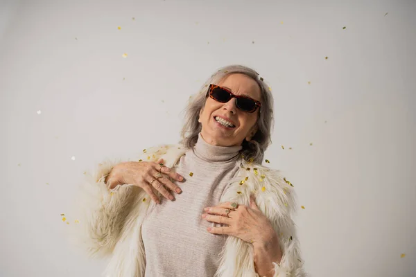 Smiling senior woman in white faux fur jacket and trendy sunglasses near falling confetti on grey background — Stock Photo