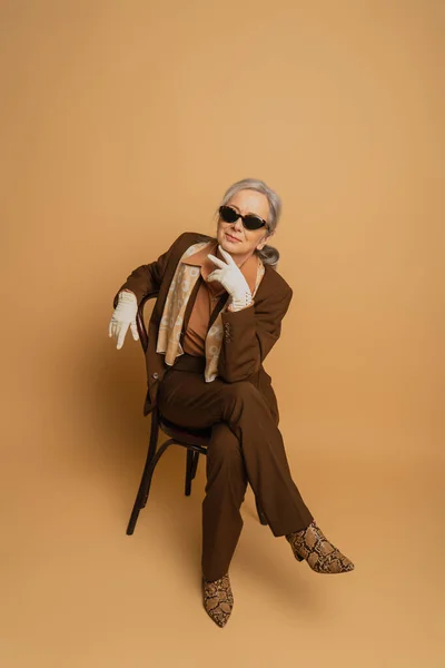 Pleine longueur de femme âgée souriante en costume marron et lunettes de soleil assis sur la chaise et regardant la caméra sur beige — Photo de stock