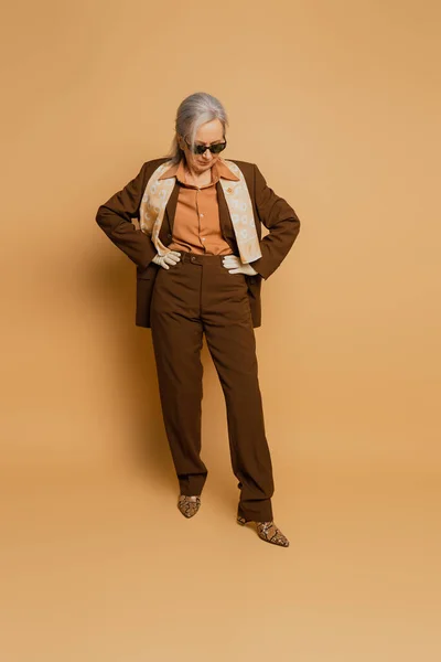 Full length of stylish senior woman in sunglasses and suit posing with hands on hips on beige — Stock Photo