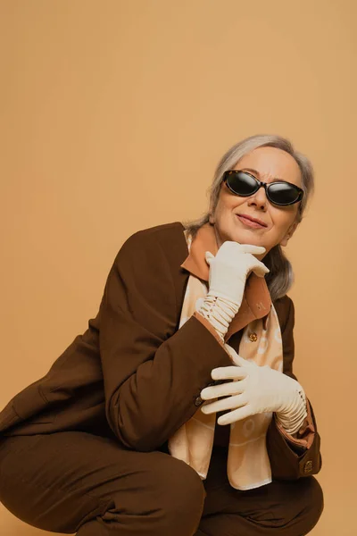 Femme âgée souriante et élégante en lunettes de soleil et costume assis isolé sur beige — Photo de stock