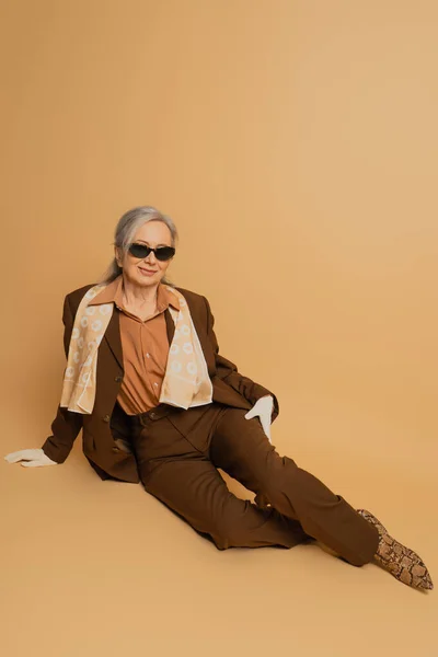 Full length of cheerful senior woman in sunglasses and suit sitting on beige background — Stock Photo