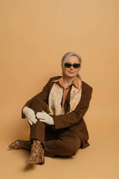 Full length of cheerful senior woman in sunglasses and suit sitting and smiling on beige background — Stock Photo