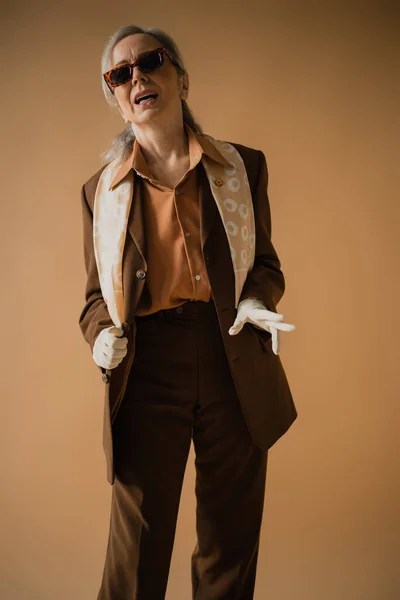 Emotional senior woman in brown suit and white gloves posing in stylish sunglasses  on beige — Stock Photo