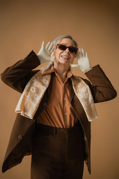 Femme âgée souriante en costume marron et gants blancs portant des lunettes de soleil élégantes sur beige — Photo de stock