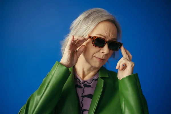 Senior woman in green jacket adjusting trendy sunglasses isolated on blue — Stock Photo