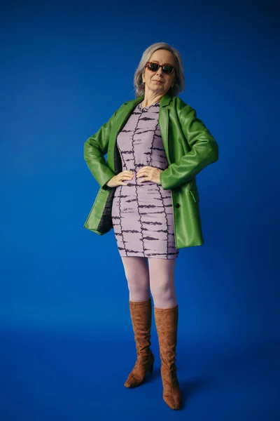 Longitud completa de la mujer mayor en chaqueta de cuero verde y vestido de pie con las manos en las caderas en azul - foto de stock
