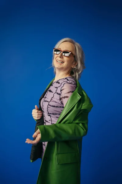 Seniorin mit Sonnenbrille und grüner Lederjacke lächelt vereinzelt auf blauem Grund — Stockfoto