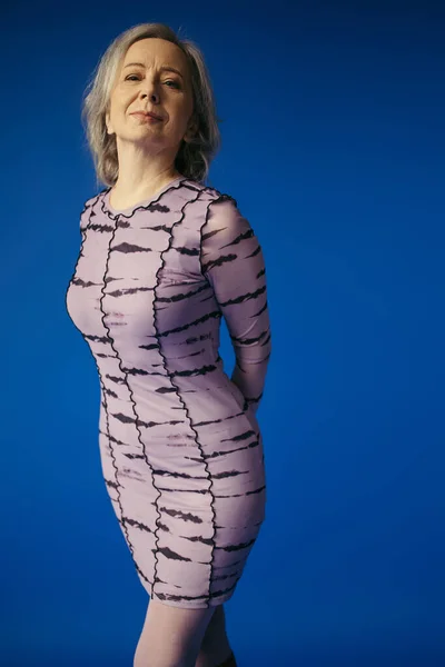 Elegant grey haired lady in purple dress posing with hands behind back and looking at camera isolated on blue — Stock Photo