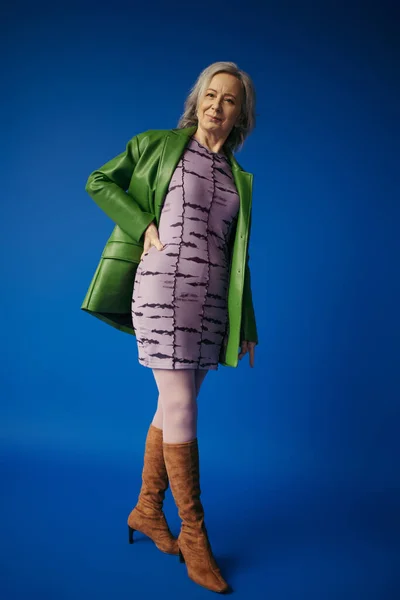 Full length of grey haired woman in trendy casual outfit standing with hand on hip on blue background — Stock Photo