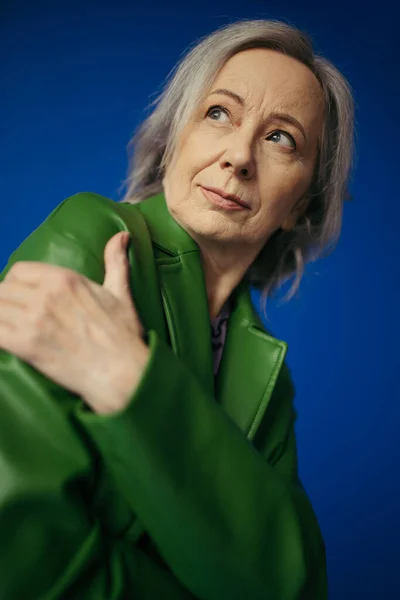 Low angle view of thoughtful grey haired woman in green leather jacket looking away isolated on blue — Stock Photo