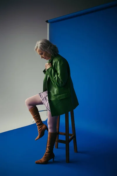 Trendy senior woman in green leather jacket and brown suede boots posing near stool on blue and grey background — Stock Photo