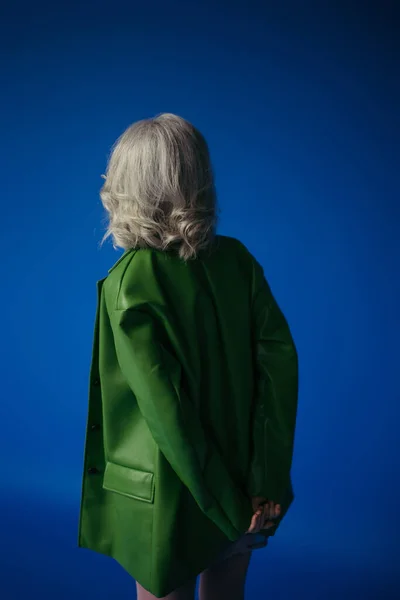 Back view of grey haired woman in green leather jacket standing with hands behind back isolated on blue — Stock Photo