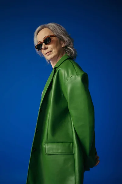 Senior model in stylish sunglasses and green leather jacket posing isolated on blue — Stock Photo