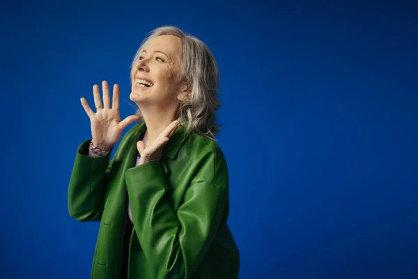 Excited grey haired woman in green leather jacket showing wow gesture and looking away isolated on blue — Stock Photo