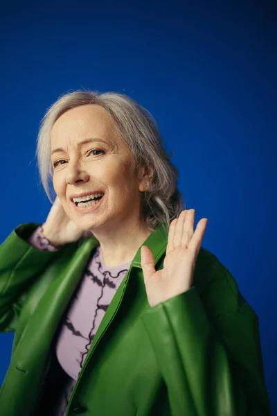 Happy grey haired woman in stylish casual clothes waving hand while looking at camera isolated on blue — Foto stock