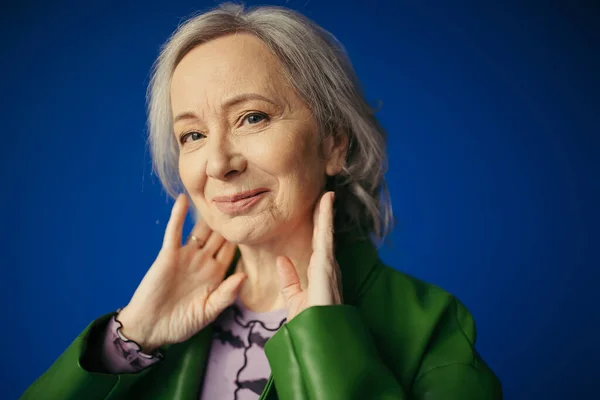 Portrait of positive and stylish senior woman posing with hands near neck and smiling at camera isolated on blue — Stock Photo