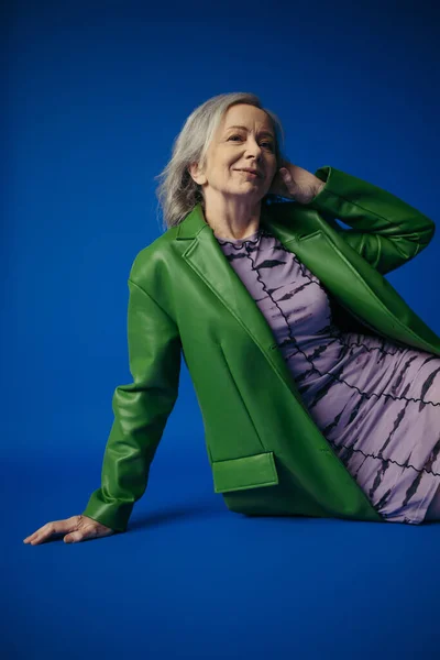 Grey haired woman in elegant lilac dress and green leather jacket smiling at camera while sitting on blue background — Stockfoto