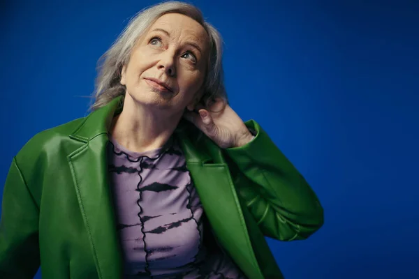 Portrait of senior model in green leather jacket and purple dress holding hand behind head and looking away isolated on blue — Stock Photo