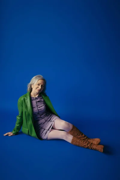 Volle Länge der glücklichen Senior-Modell in stilvoller Freizeitkleidung sitzt auf blauem Hintergrund — Stockfoto