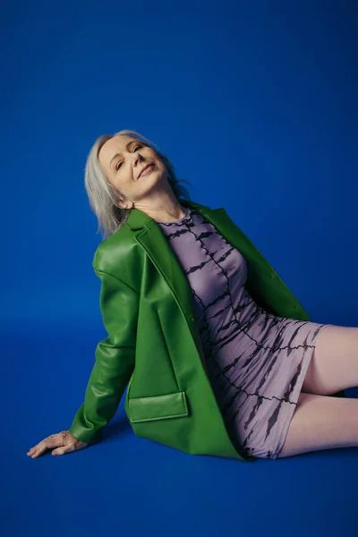 Happy grey haired woman in green leather jacket and purple dress looking at camera while sitting on blue background — Stockfoto
