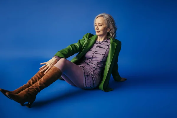 Full length of trendy senior woman in suede boots and green leather jacket over lilac dress sitting on blue background — Photo de stock